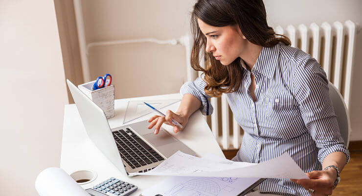 Une jeune femme est assise à un bureau, devant un écran d’ordinateur, avec autour d’elle des documents, des stylos et une calculette